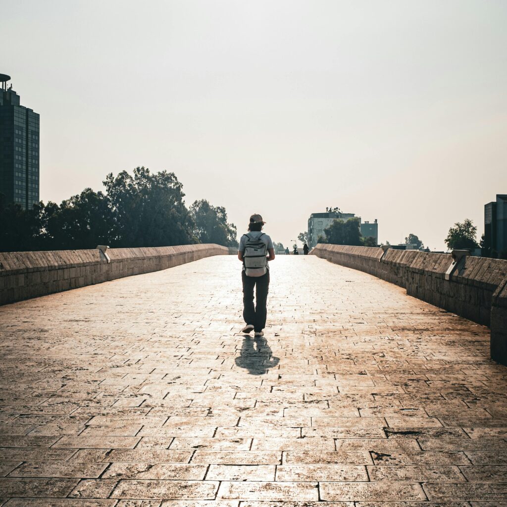 A lone traveler walks on a sunlit stone bridge in an urban setting, capturing adventure.